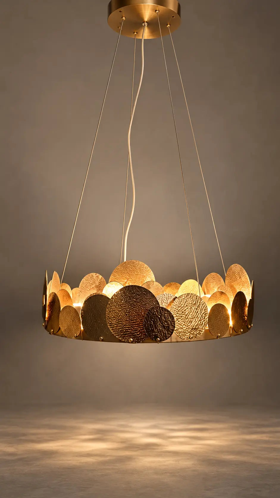 A modern circular pendant chandelier with a brushed gold and textured bronze metallic disc design, suspended by thin cables against a warm gray background.