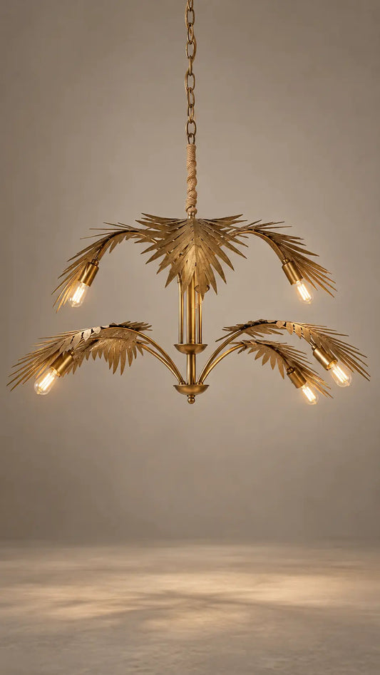 A gold-finished palm tree chandelier with multiple leaf-shaped arms and glowing light bulbs, suspended from a chain against a neutral gray studio background.